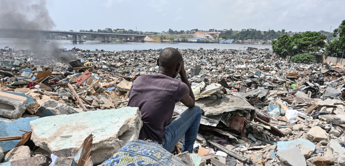 man in Ivory Coast sitting in front of a destroyed village