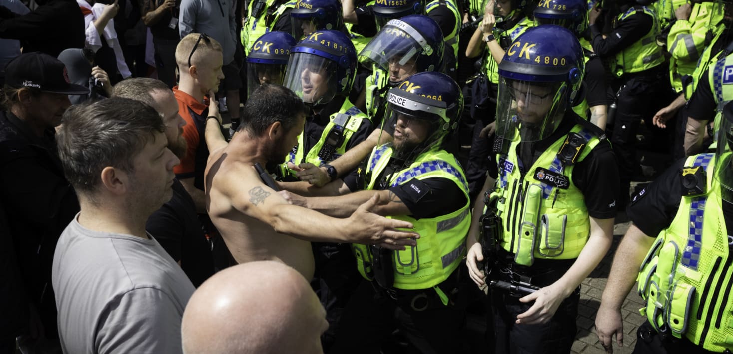 police in yellow vests keeping a man without a shirt from moving forward.